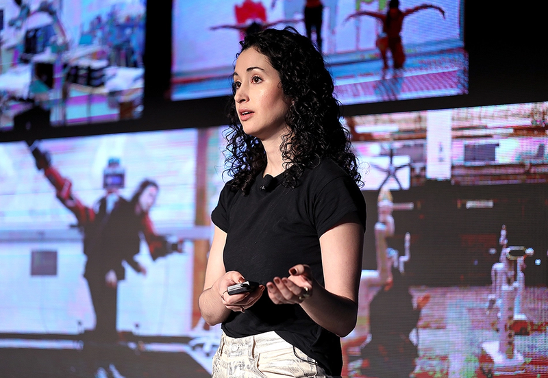 A woman presenting on stage in front of a screen showing motion capture visuals