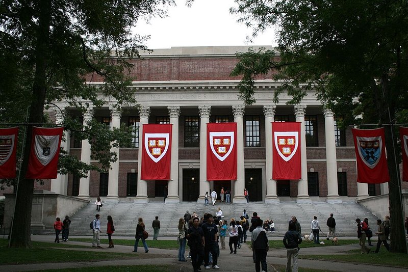 A crowd of students walk in front of a Harvard University library, which has the institution's seal on red flags hanging from it.