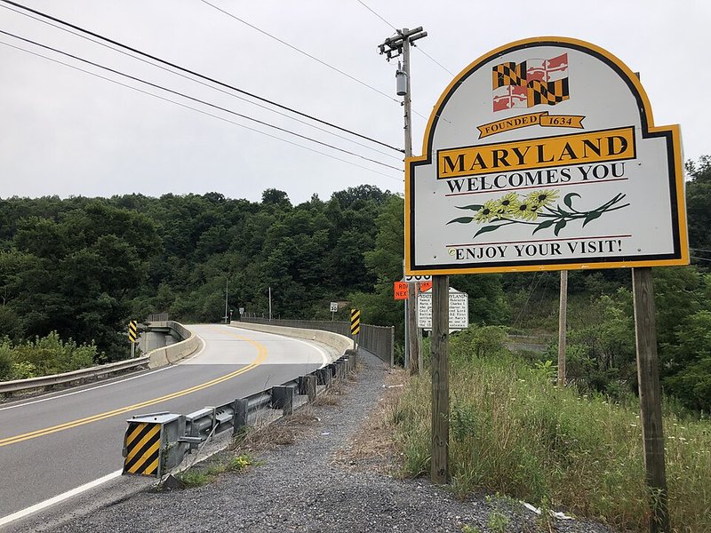 A white, yellow, and black sign that says "Maryland Welcomes You" on the side of an empty road curving towards woods.