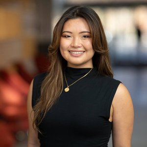 Gracielle Li with long, wavy brown hair, smiling at the camera. She is wearing a sleeveless black top and a gold necklace with a round pendant. The background is softly blurred, suggesting an indoor setting with warm lighting