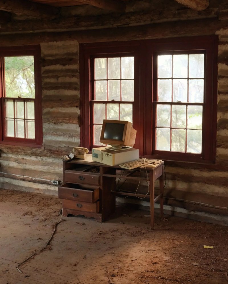 An old computer and rotary phone sit on a dusty wooden desk inside a vacant school building, symbolizing decline and abandonment in small college towns.
