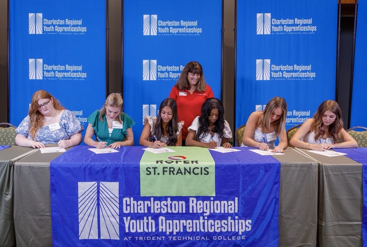 Six youth apprentices sitting down at a table signing their contract with an employer behind them.