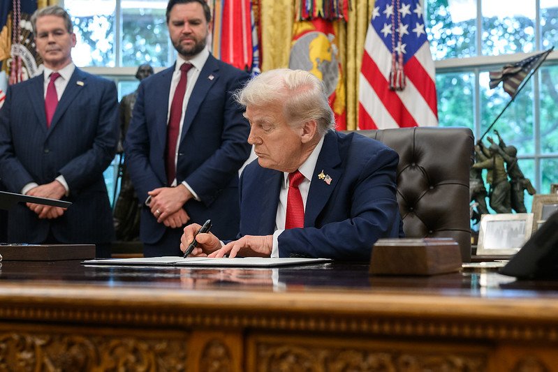 President Trump sits in Oval Office signing official documents with Vice President JD Vance and others in the background