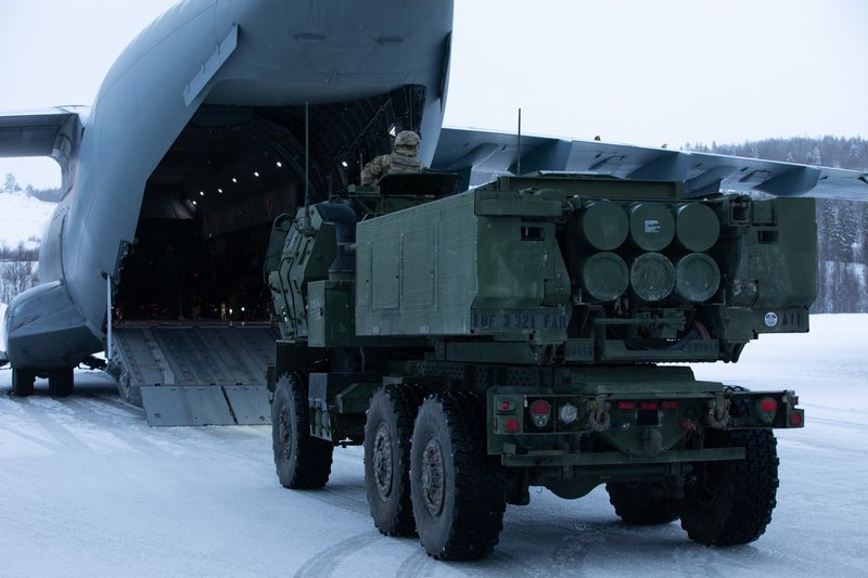 Military vehicle carrying missiles being unloaded from a transport aircraft on a runway.