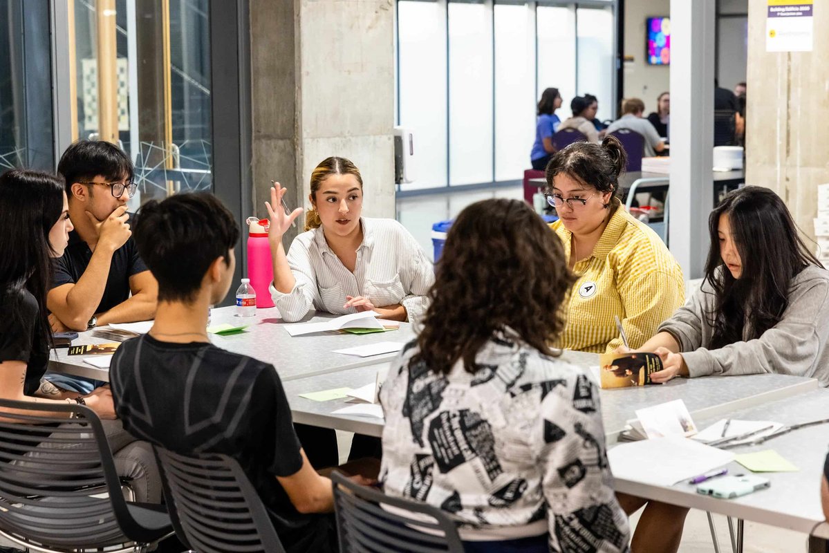 A group of young adults sitting around a table in discussion, with papers and pens present.