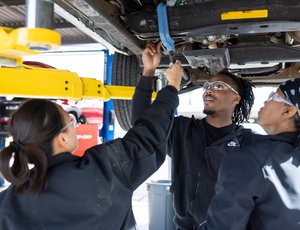 Multiethnic group of students in an automotive technology program working on the undercarriage of a pickup truck at an outdoor auto shop on a college campus.