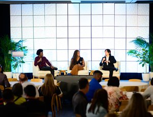 Three youth apprentices speak on a panel at a conference. They sit in chairs on a stage while audience members listen.