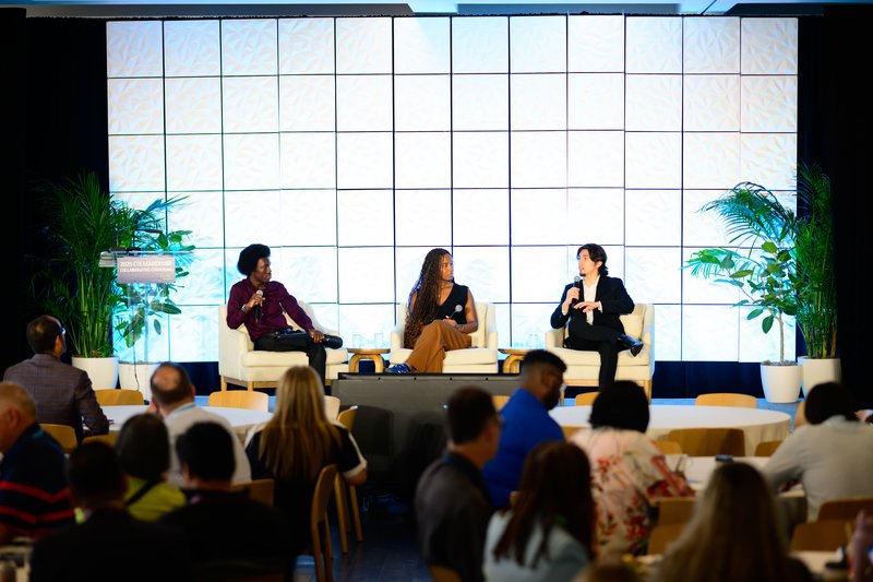 Three youth apprentices speak on a panel at a conference. They sit in chairs on a stage while audience members listen.