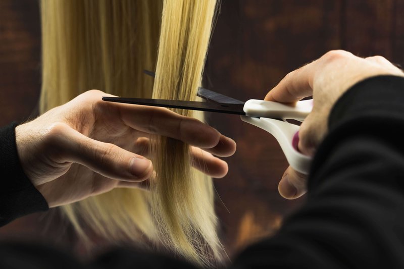 A close-up image of hands holding a long strand of blonde hair that it is about to cut with scissors.