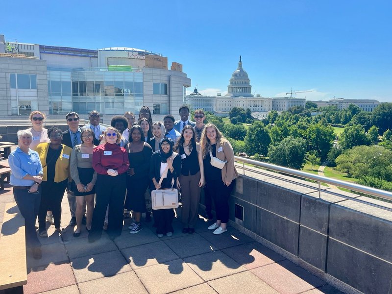 The PAYA Youth Council, a group of 15 young adults, poses for a group photo on the roof of the U.S. Department of Labor. The dome of the Capitol building is visible in the background.