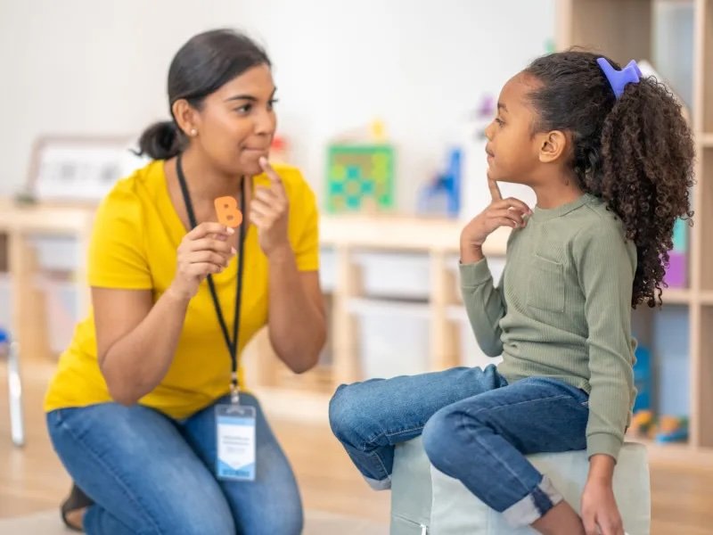 A speech therapist kneels in front of a young girl in a classroom