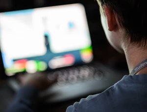 A teenager with a lanyard sits in front of a laptop in a dimly lit room.