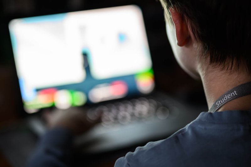 A teenager with a lanyard sits in front of a laptop in a dimly lit room.