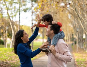 A joyful family moment with a man carrying a child on his shoulders, reaching out to high-five a woman in a park setting.