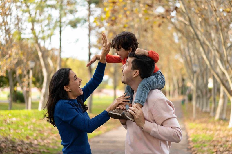A joyful family moment with a man carrying a child on his shoulders, reaching out to high-five a woman in a park setting.