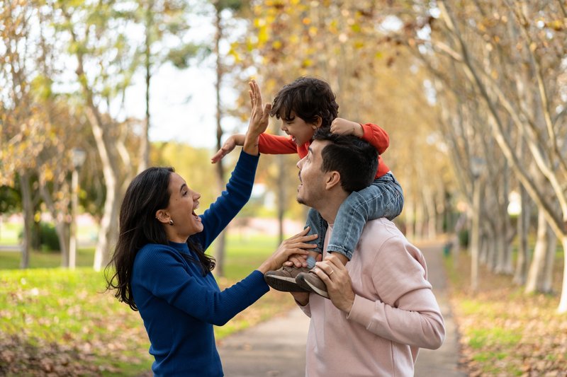 A joyful family moment with a man carrying a child on his shoulders, reaching out to high-five a woman in a park setting.