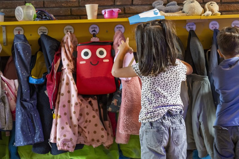 A small child faces a wall of jacket hooks, from which kids' coats and a red smiley face backpack hangs.