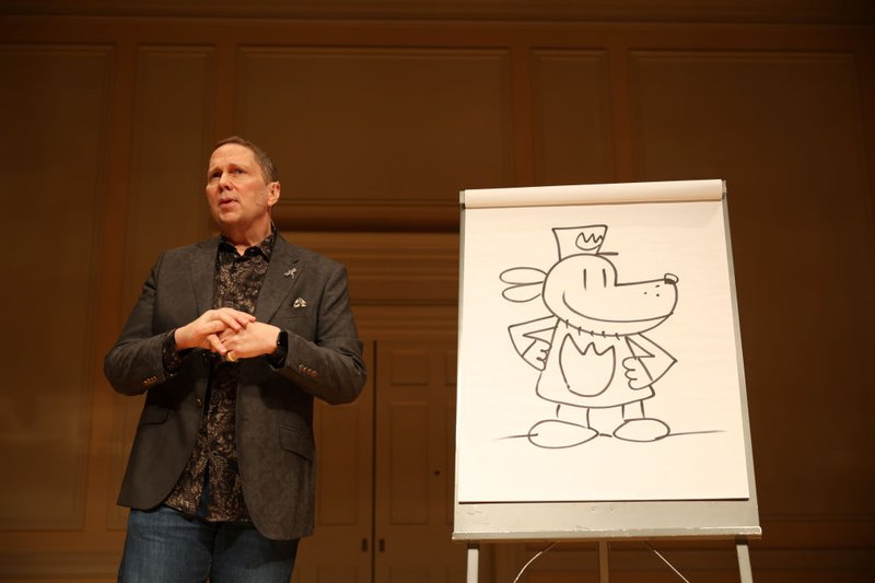 Dav Pilkey speaks onstage in front of a Dog Man drawing during a special reading event.