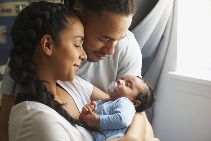A young woman holds a sleeping newborn with a pacifier in his mouth while a taller man stands beside the woman with his arms wrapped around both the woman and child.