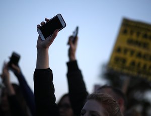 People raising smartphones in the air, with a blurred protest sign in the background.