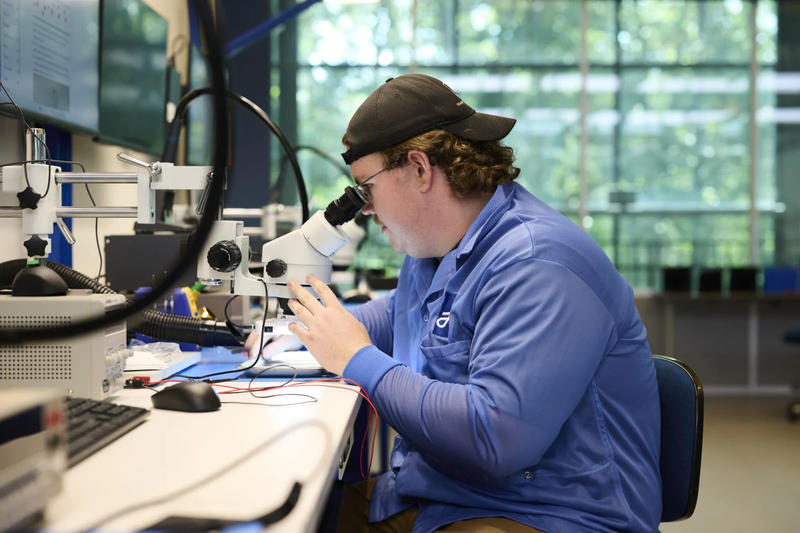A student in a blue lab coat and backwards cap looks through a microscope while working with electronic components in a college manufacturing training lab.