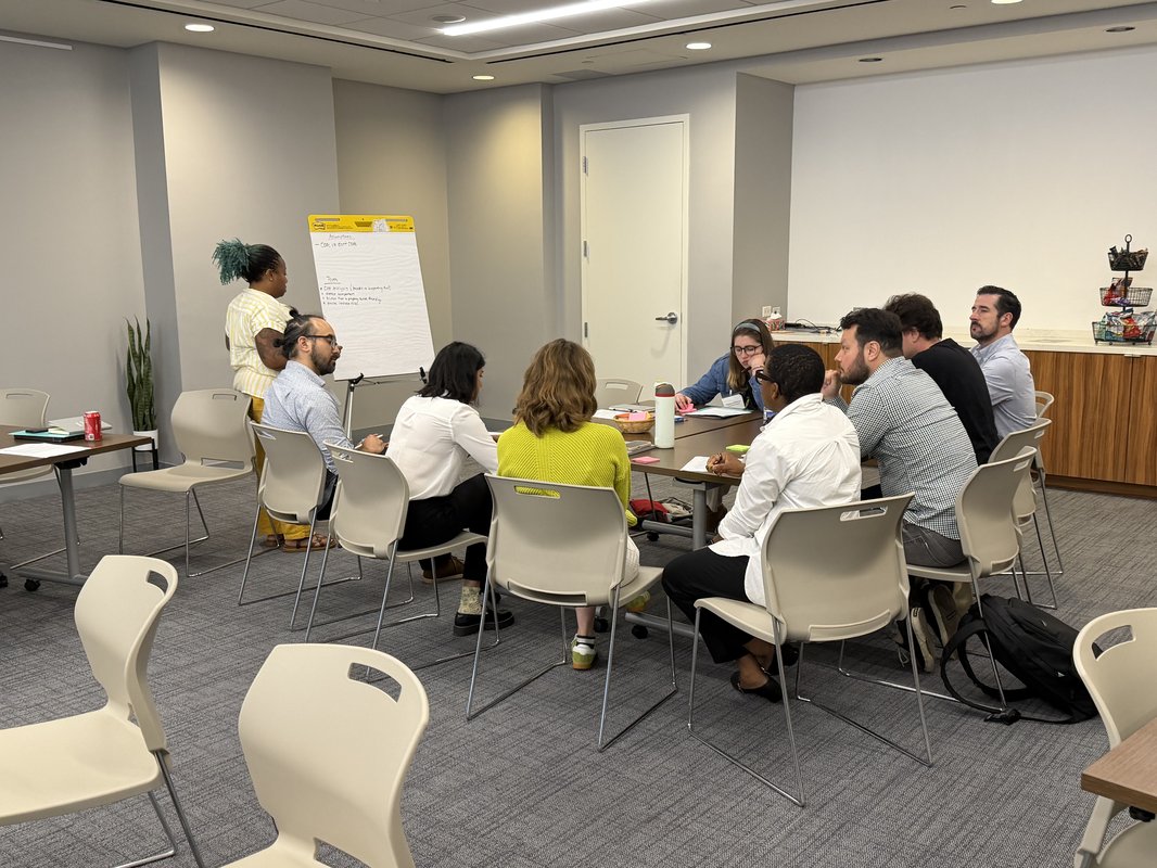 Nine people gathered around a table, with one person taking notes on a flipchart.