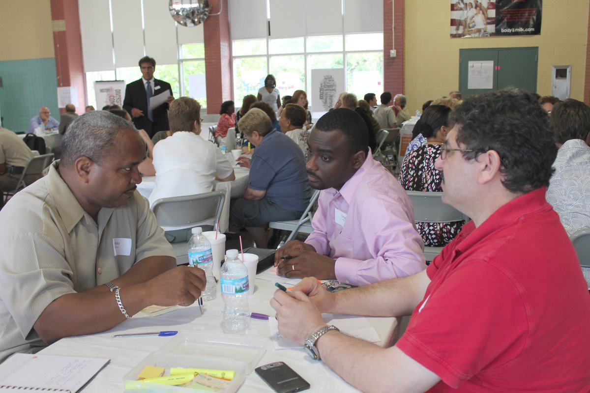 Three men in business casual attire sit at a table with water bottles and post-it notes on it. In the background are several other tables with similar groups of adults having conversations.