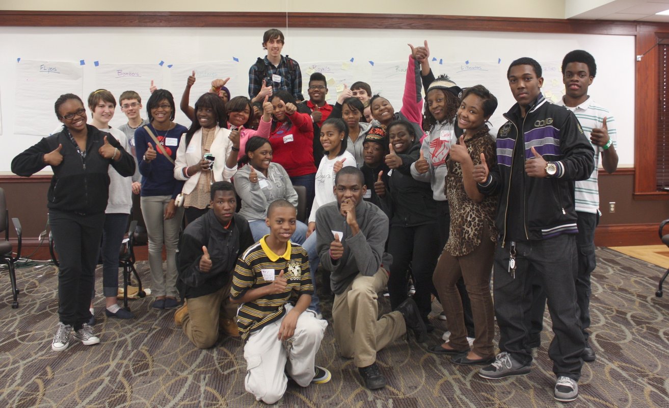 About twenty high-school students pose for a group photo in front of a white board. Many give thumbs-ups.