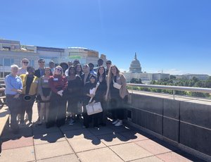 The PAYA Youth Council, a group of 15 young adults, poses for a group photo on the roof of the U.S. Department of Labor. The dome of the Capitol building is visible in the background.