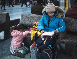A student parent sits in a public space reviewing papers while a young child plays beside them.