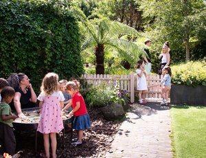 Several children and an adults are gathered around a water table in a lush garden, while other adults chat near a white picket fence under bright sunlight.