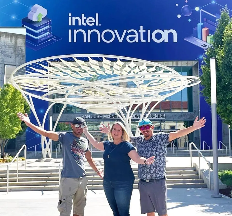 Three individuals stand in front of a building labeled "Intel Innovation," with their arms outstretched.
