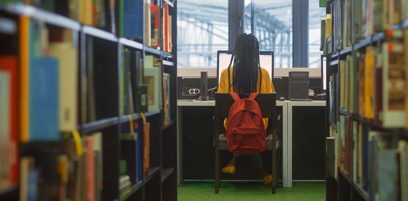 Student on a computer at the library surrounded by stacks of books.