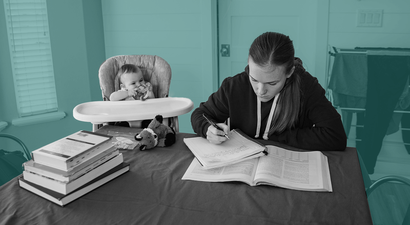 A woman sitting next to infant in high chair takes notes at table with pile of books, against teal backdrop.