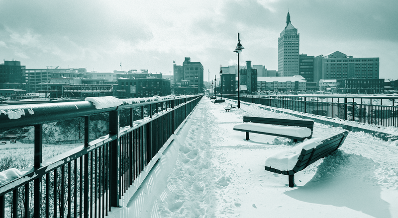 A snow-covered bridge with benches crosses toward a city skyline, with a teal overlay.