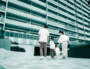 Two parents hold the hands of their two small children outside entrance of large apartment building, with a teal overlay.