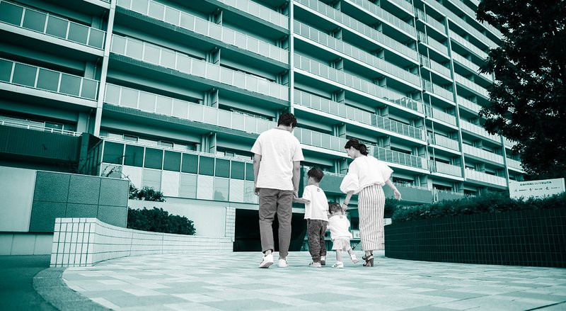 Two parents hold the hands of their two small children outside entrance of large apartment building, with a teal overlay.