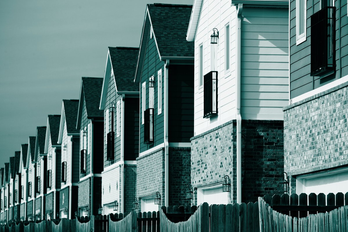 A row of narrow, similar-looking single family homes behind wooden fences, in teal.