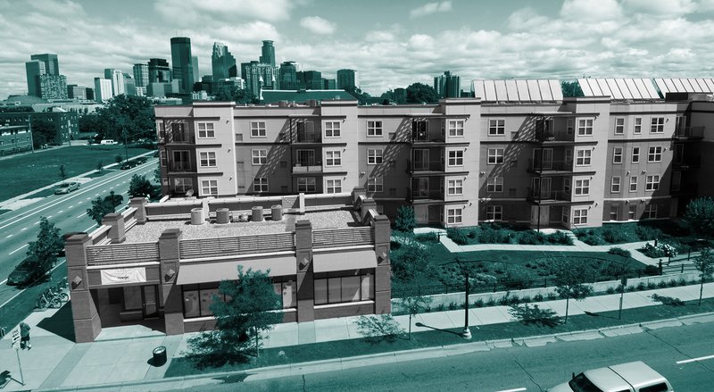 An apartment complex sits behind a smaller building, sidewalk, and street in front of a city skyline, with teal overlay.