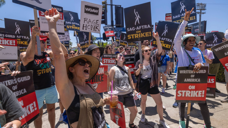 A crowd of SAG‑AFTRA and Writers Guild of America members hold strike signs and chant during a lively picket line, calling for fair contracts and protections, including around AI use in entertainment.