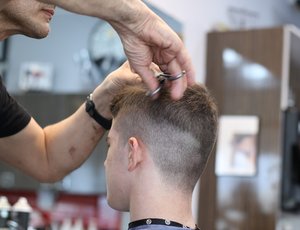 The back of a person's head is visible as they get their hair cut short. The lower half of the stylist's face, plus their arms and hands, are visible.