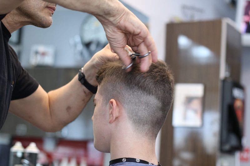 The back of a person's head is visible as they get their hair cut short. The lower half of the stylist's face, plus their arms and hands, are visible.