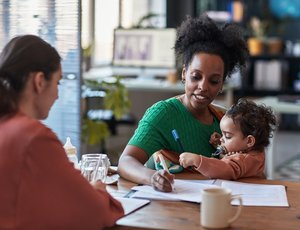 a black mom is holding her baby and trying to work at a desk facing another woman