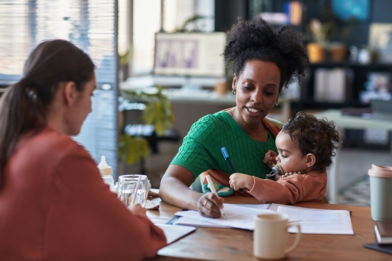 a black mom is holding her baby and trying to work at a desk facing another woman
