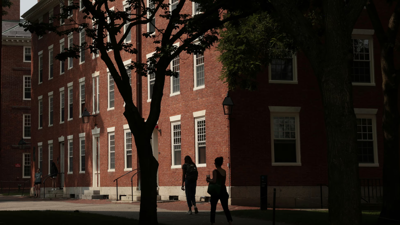 A student walks past the main entrance of a university campus lined with American flags. The image evokes the intersection of higher education and national politics central to debates over Trump’s proposed “Compact for Academic Excellence.”