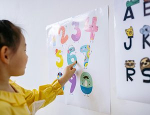 A young girl points to cartoon numbers on a poster on the wall.