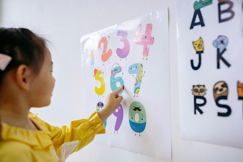 A young girl points to cartoon numbers on a poster on the wall.