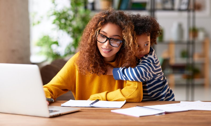 Parenting student wearing glasses using a laptop and thinking about work task while a small boy son gently hugs her.
