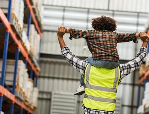 Person wearing a high-visibility vest carries a child on their shoulders in a warehouse aisle.