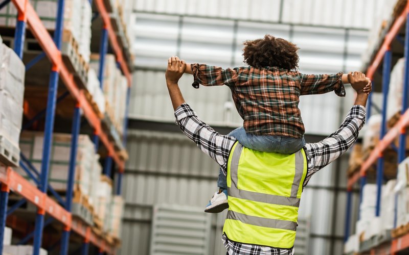 Person wearing a high-visibility vest carries a child on their shoulders in a warehouse aisle.